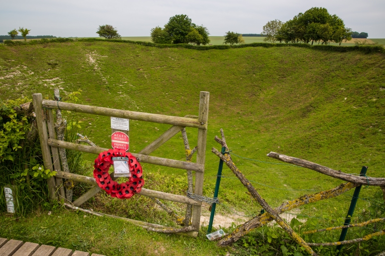 A wreath of poppies hangs on a wooden gate, beyond which lies the Lochnagar Crater